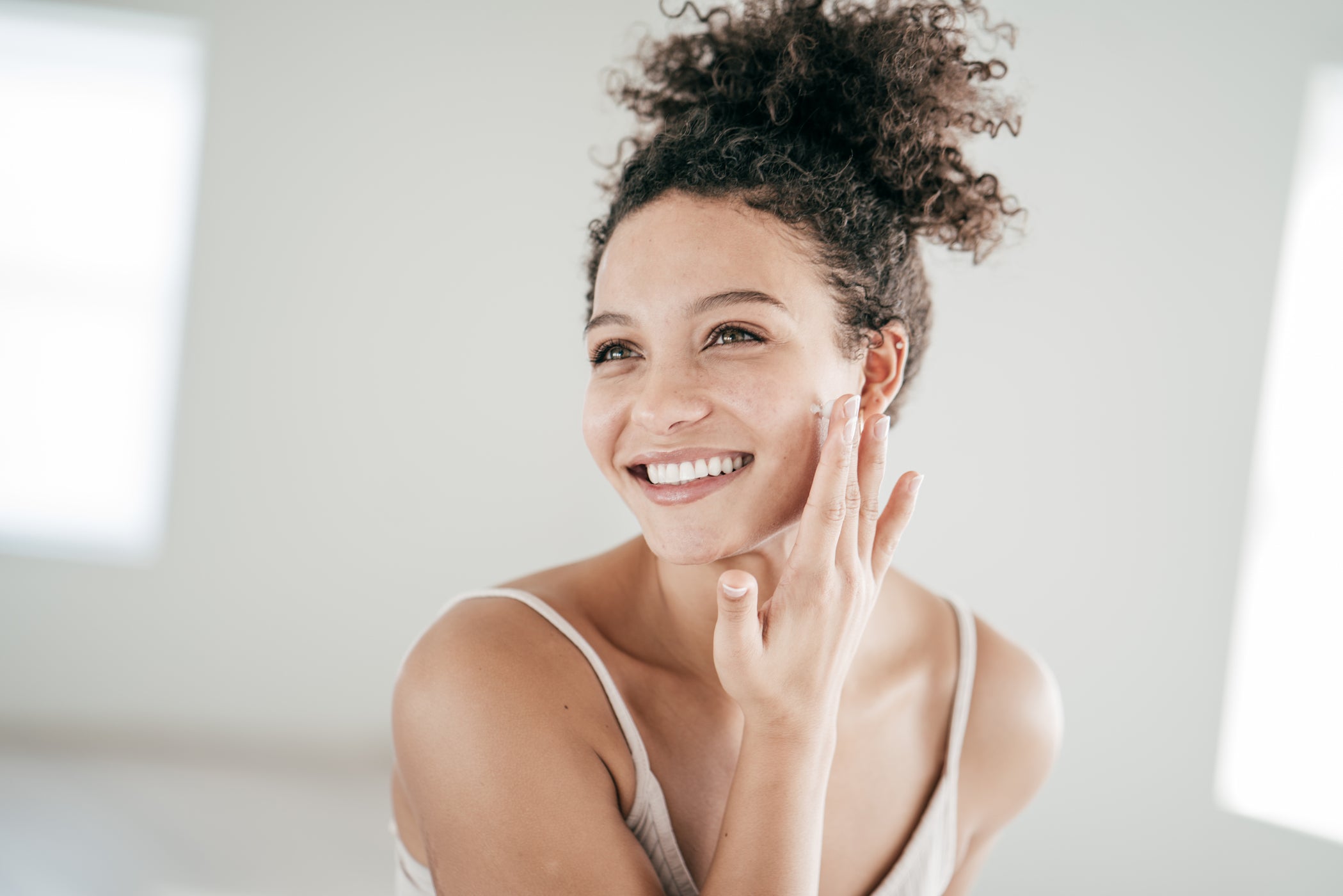Jeune femme aux cheveux foncés et bouclés sourit et s'applique de la crème sur les joues avec le doigt