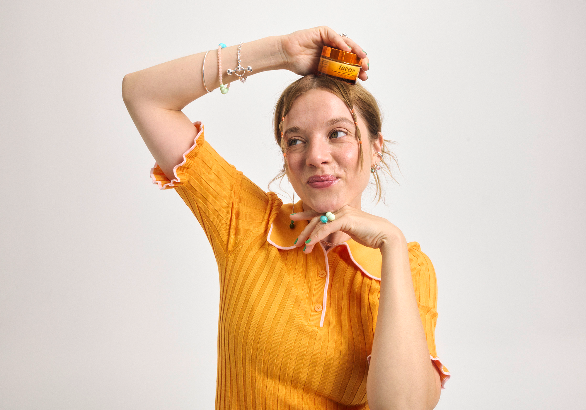 Jella Haare in orangefarbenem Shirt hält spielerisch ein lavera Glow Produkt über ihren Kopf.