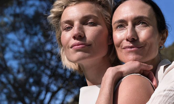 Una modella bionda e una dai capelli scuri stanno davanti a un albero e si tengono strette.