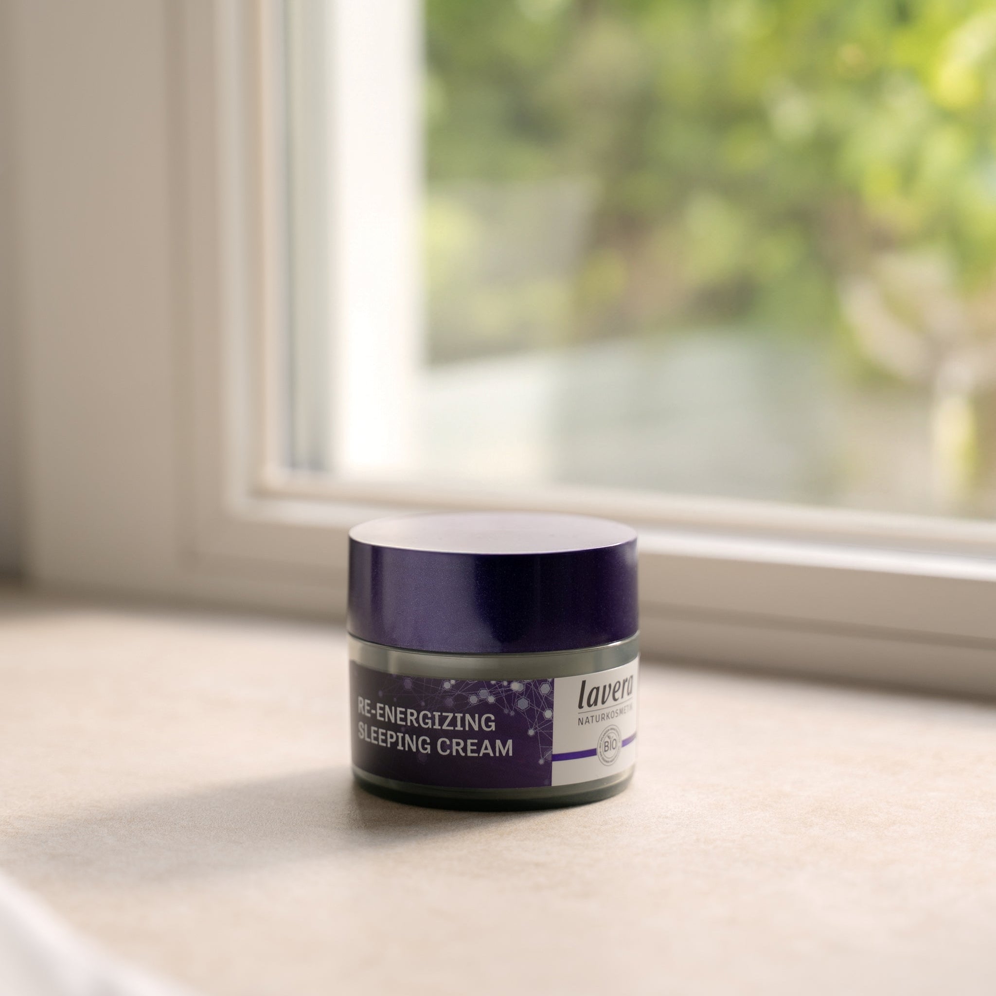 Un barattolo rotondo con coperchio viola del prodotto lavera Naturkosmetik Re-Energizing Sleeping Cream, posizionato su un davanzale alla luce soffusa del giorno, con sfondo verde sfocato.